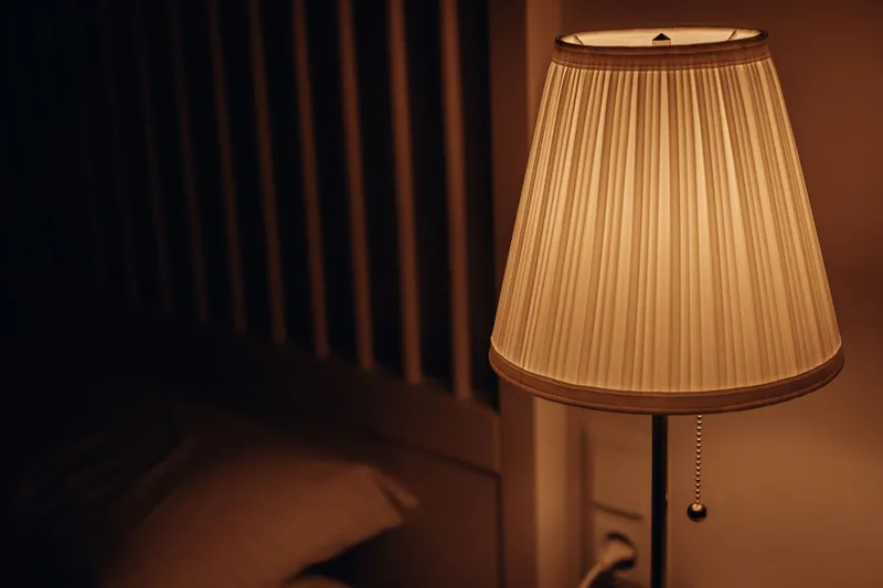 A pleated bedside lamp glowing warmly beside a dark bed at night