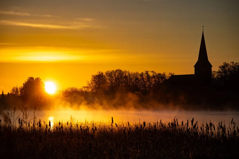 Golden sunrise over a misty lake, with tall reeds in the foreground and the silhouette of a village church spire rising above the trees on the far bank