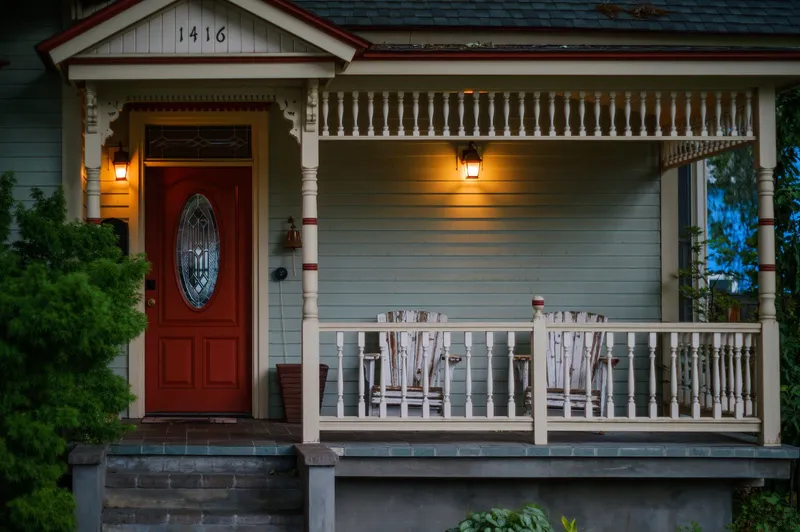A wide front porch at dusk with two empty white rocking chairs, warm lamps lit on either side of a red front door