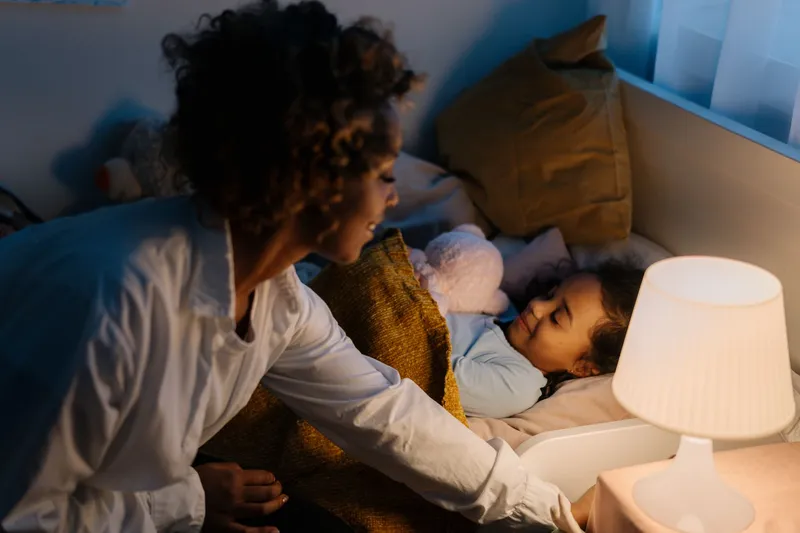 A mother leaning over her young daughter to tuck her in, lit by a small bedside lamp in a dim bedroom