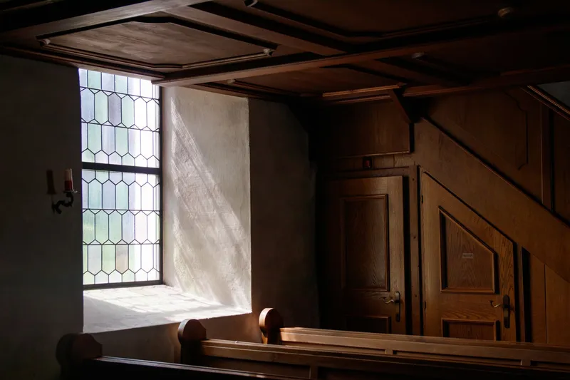 Morning light falling through a leaded glass window onto the wall of a quiet church, above empty wooden pews